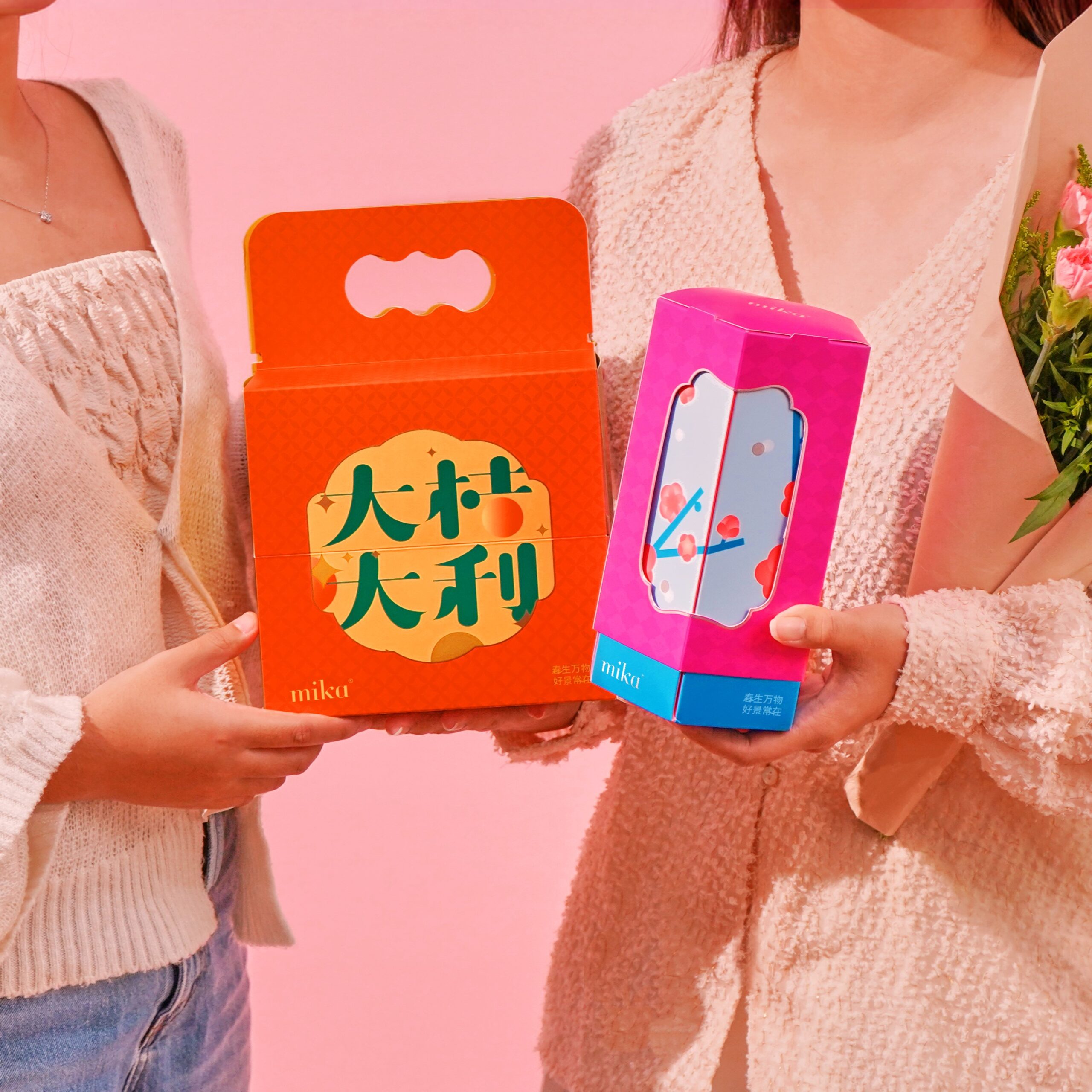 Two people in soft sweaters hold bright orange and pink gift boxes against a pink background, front view.